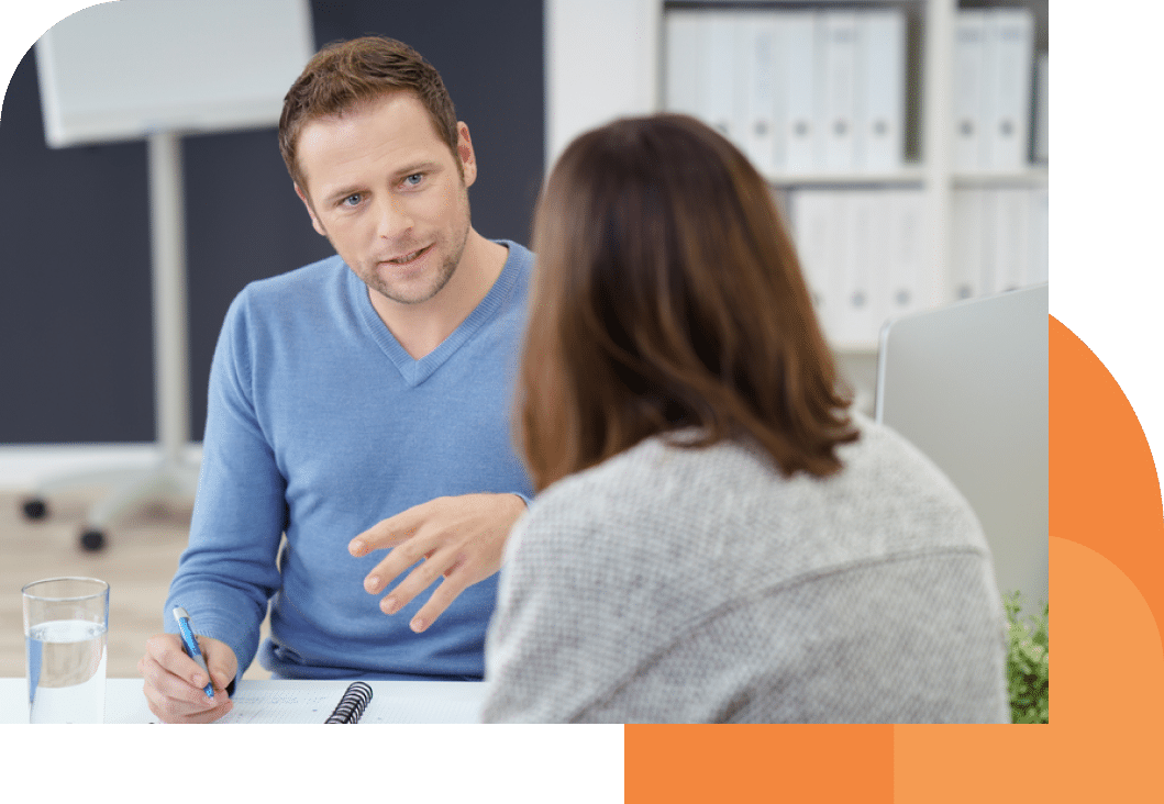 This photo shows a man facing the camera, talking to a woman who has her back to the camera. They are sitting at a table and the man has a pen in his hand. It looks like he's in mind sentence. This image depicts a mortgage advisor talking to a client.