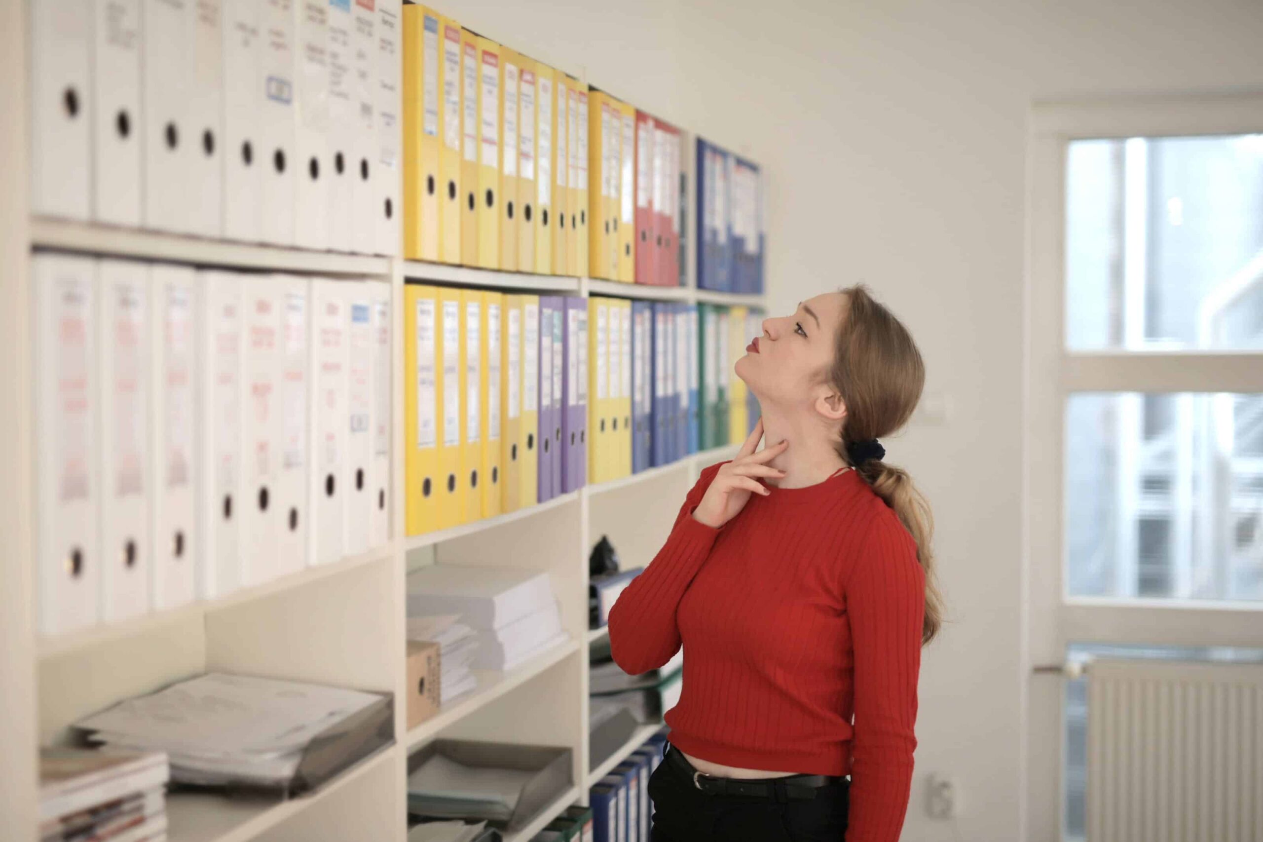 Lady wearing a red jumper is looking at a wall on which lots of level arch files are stored. This depicts the manual process that Gatheroo replaces with digital file collection, client collaboration and automated client reminders.