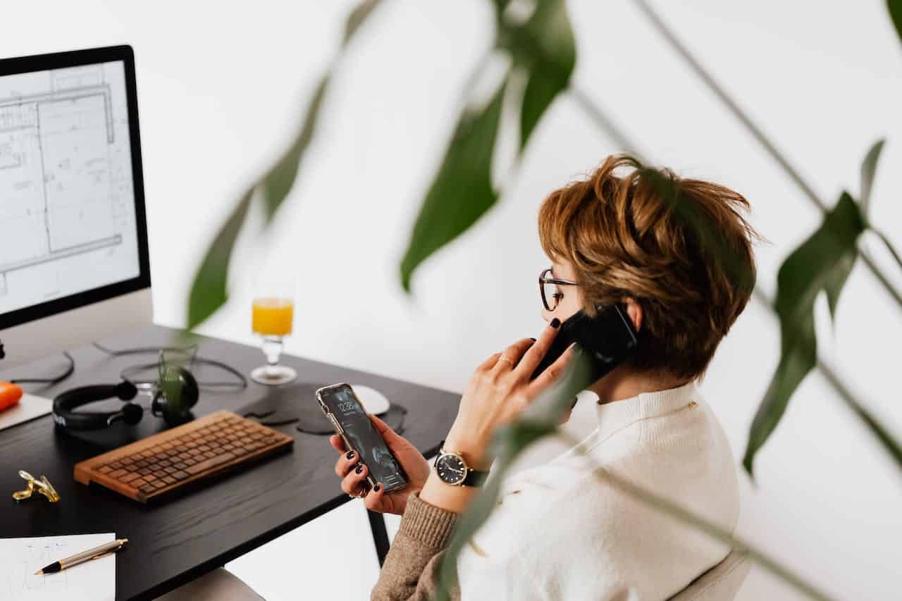 This photo shows a lady sitting at her desk with a phone in one hand at her ear, and looking at a mobile phone in the other hand, seemingly communicating with clients.